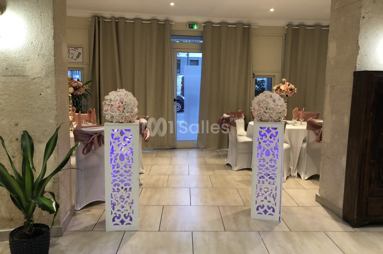 Salle décorée pour un événement, avec tables ornées de fleurs et colonnes blanches ajourées devant des rideaux beiges.