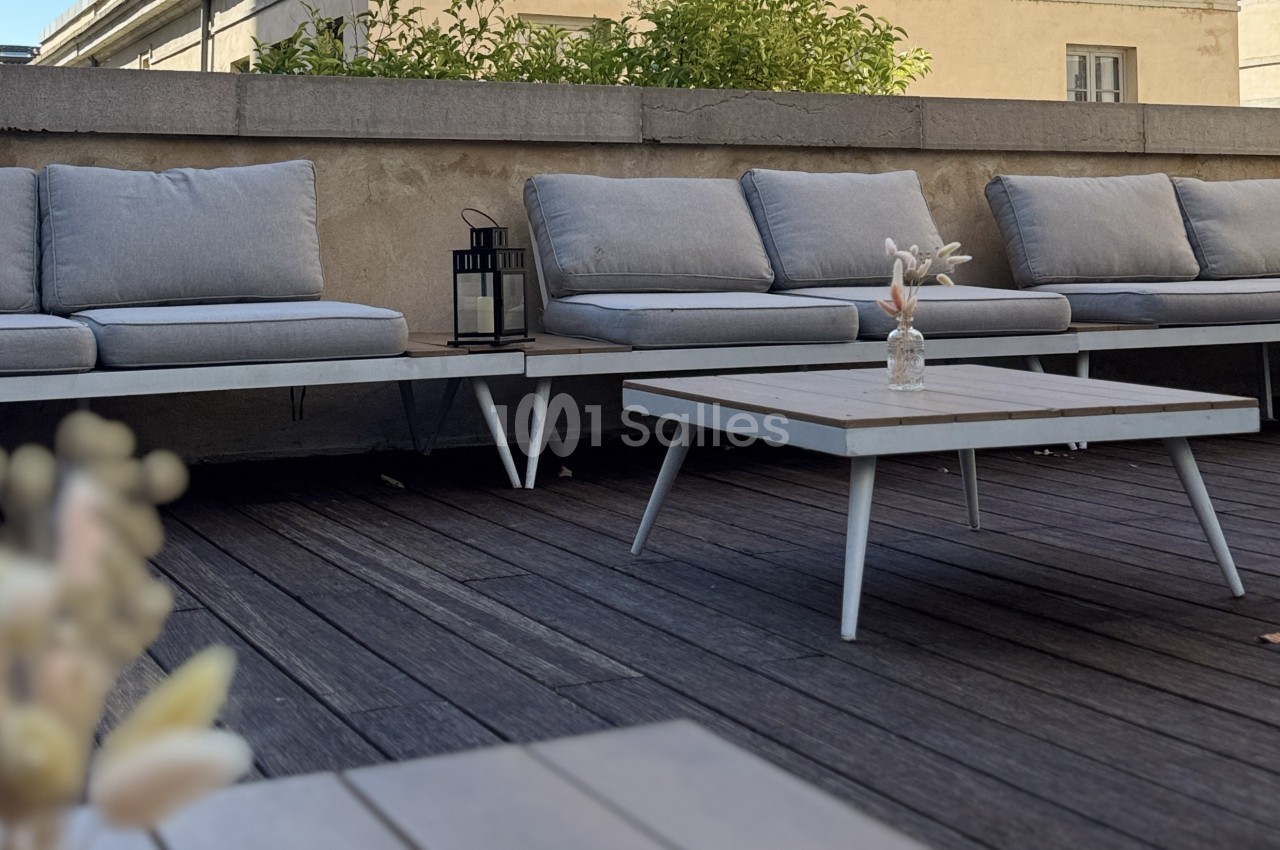 Bancs gris avec coussins et table basse en bois sur une terrasse en bois, décorée d'une lanterne et d'un vase minimaliste.