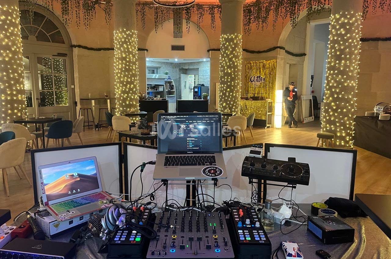 Poste de DJ avec table de mixage, ordinateurs et contrôleurs, installé dans une salle décorée de guirlandes lumineuses.