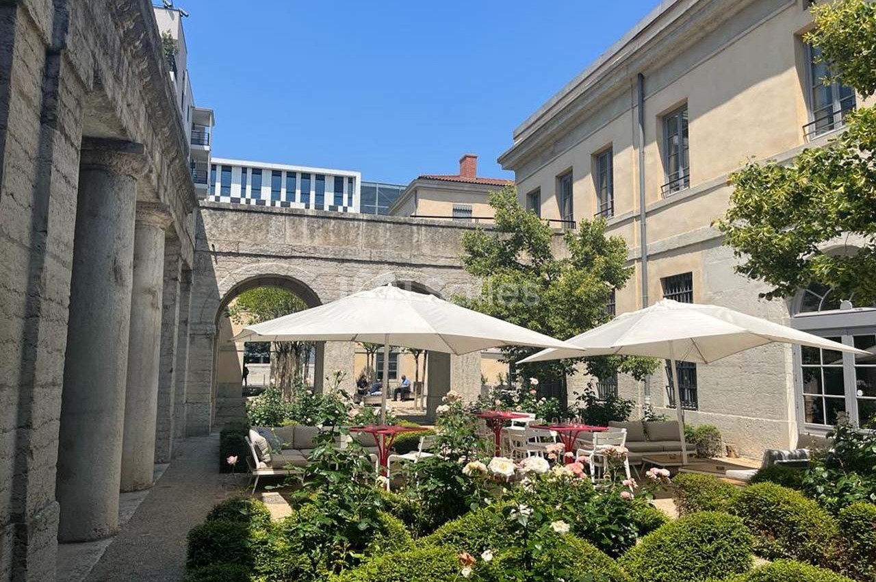 Cour intérieure avec jardin fleuri, tables et chaises sous des parasols blancs, entourée de bâtiments en pierre.