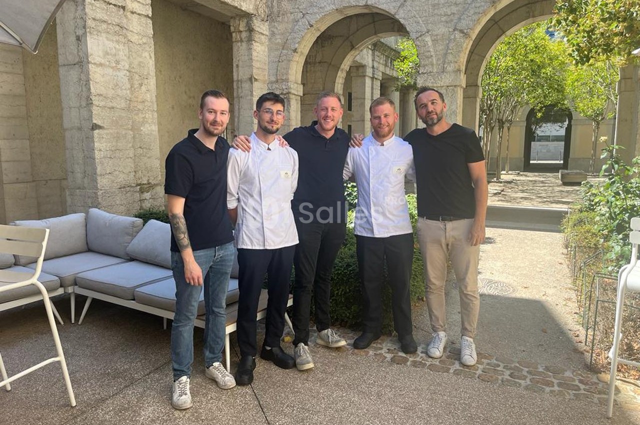 Cinq hommes posent ensemble dans une cour ensoleillée avec des arches en pierre et des meubles d'extérieur.