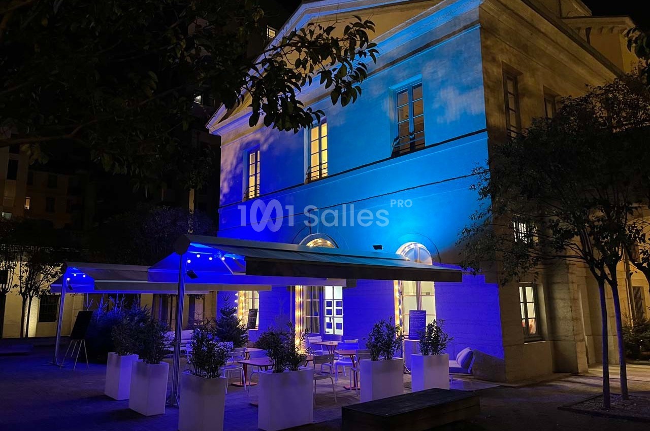 Bâtiment éclairé en bleu la nuit, avec une terrasse aménagée de tables, chaises et plantes en pots.