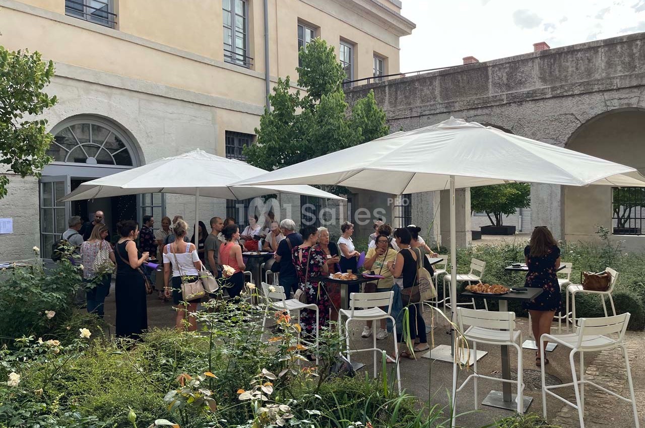 Groupe de personnes rassemblées autour de tables sous des parasols dans une cour extérieure avec végétation.