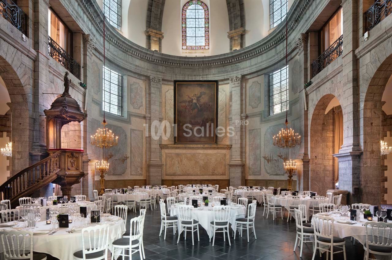 Salle de réception dans une église rénovée, avec tables rondes dressées, chaises blanches et lustres suspendus.
