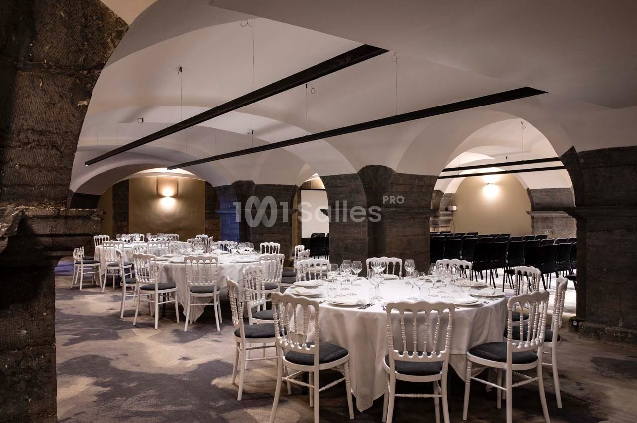 Salle voûtée avec tables rondes dressées pour un événement, chaises blanches et éclairage tamisé.