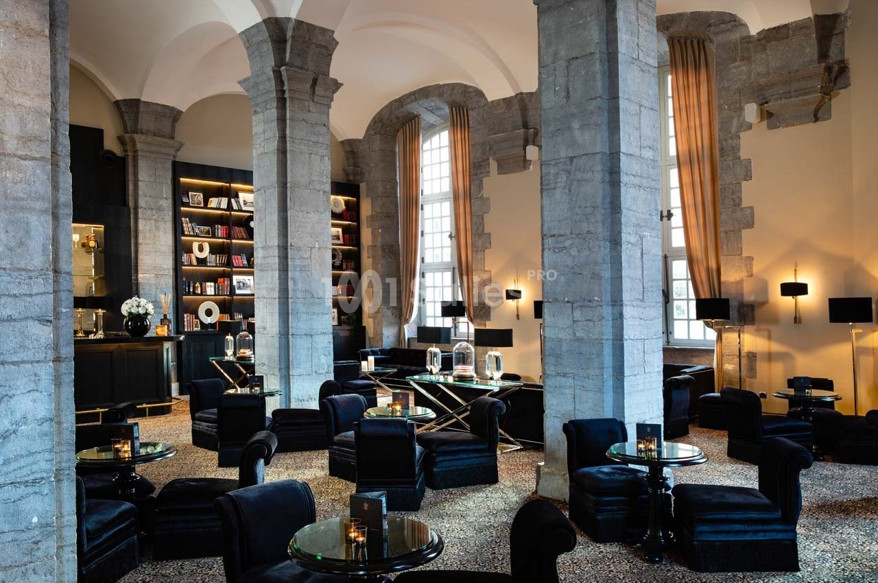 Salon élégant avec colonnes en pierre, fauteuils noirs, tables en verre et étagères remplies de livres.
