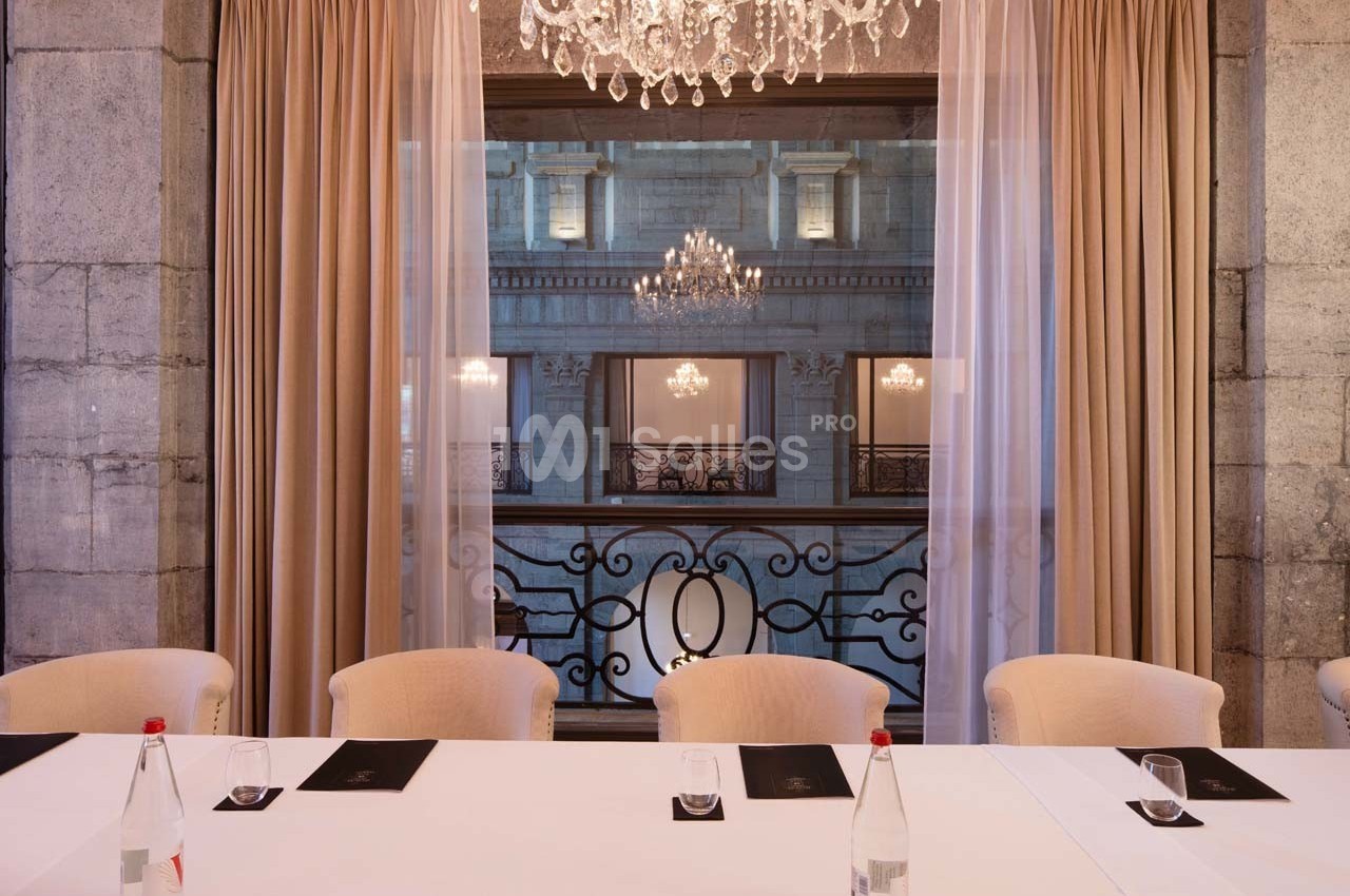 Salle élégante avec table blanche, chaises beiges, rideaux ouverts et vue sur un bâtiment illuminé par des lustres.