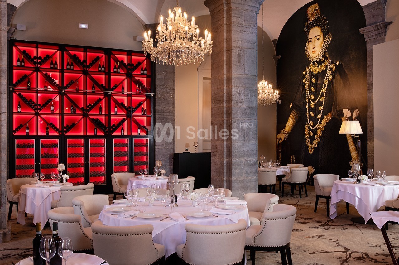 Salle de restaurant élégante avec tables dressées, lustres en cristal, mur rouge à bouteilles et fresque murale ornée.