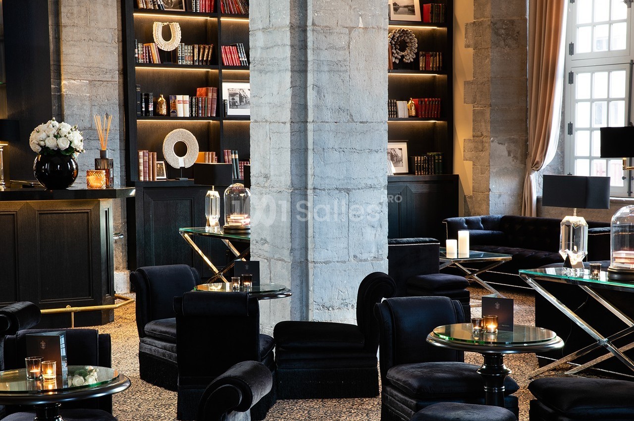 Salon élégant avec fauteuils noirs, tables en verre, étagères décorées de livres et objets, éclairage tamisé.