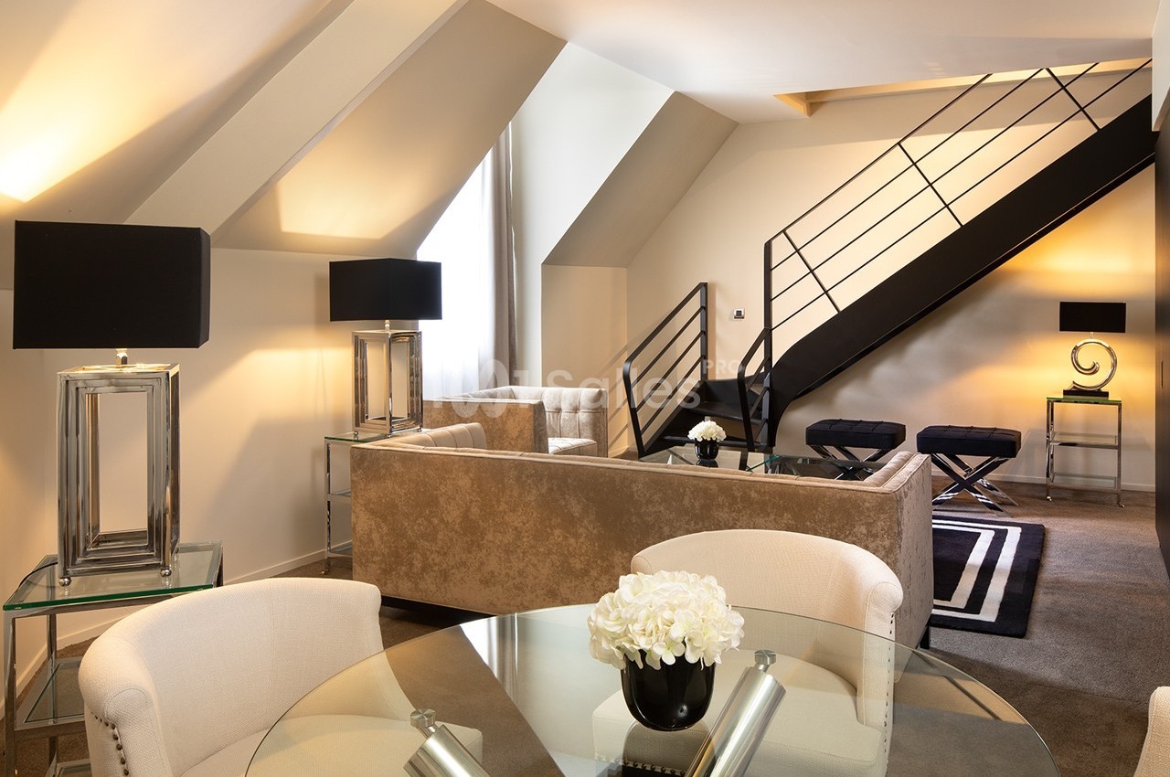 Salon moderne sous combles avec canapé beige, table en verre, lampes noires et escalier menant à une mezzanine.