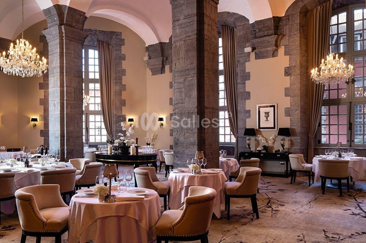 Salle de restaurant élégante avec hauts plafonds, lustres, tables rondes dressées et grandes fenêtres.