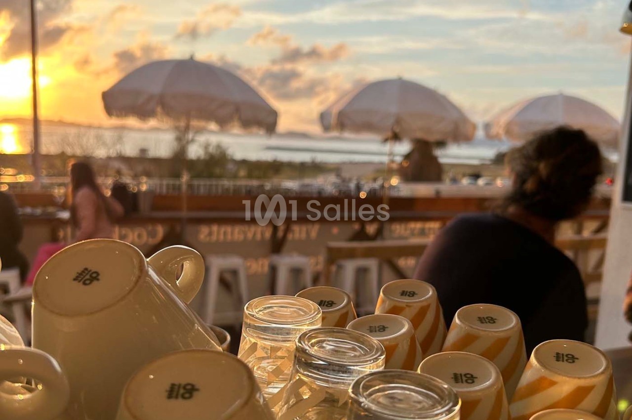 Tasses empilées et verres sur un comptoir en terrasse, avec vue sur un coucher de soleil et des parasols en arrière-plan.