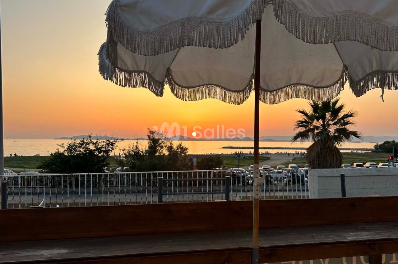 Vue d'un coucher de soleil sur la mer, avec un parasol blanc au premier plan et des palmiers en arrière-plan.
