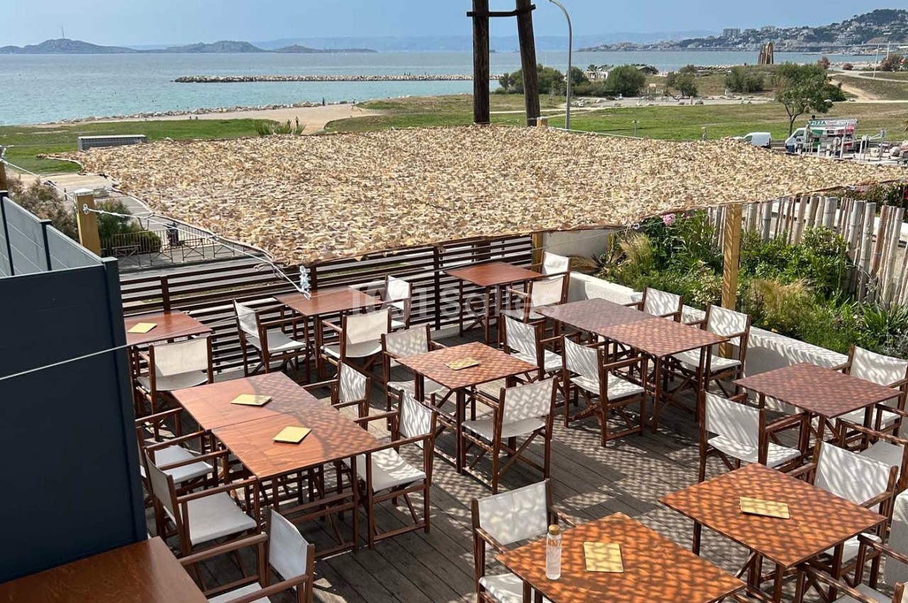Terrasse en bois avec tables et chaises, ombragée par une pergola, offrant une vue sur la mer et un paysage côtier.