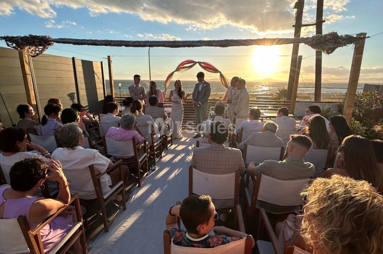 Cérémonie de mariage en extérieur au coucher du soleil, avec des invités assis face aux mariés sous une arche décorée.