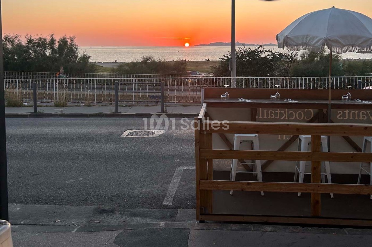 Vue d'une terrasse en bord de route avec des tabourets, face à un coucher de soleil sur la mer.