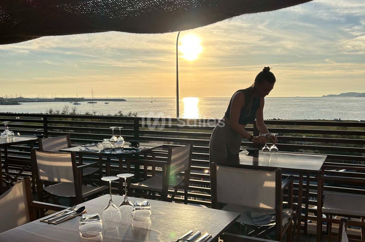 Une serveuse prépare une table en terrasse avec vue sur la mer au coucher du soleil.