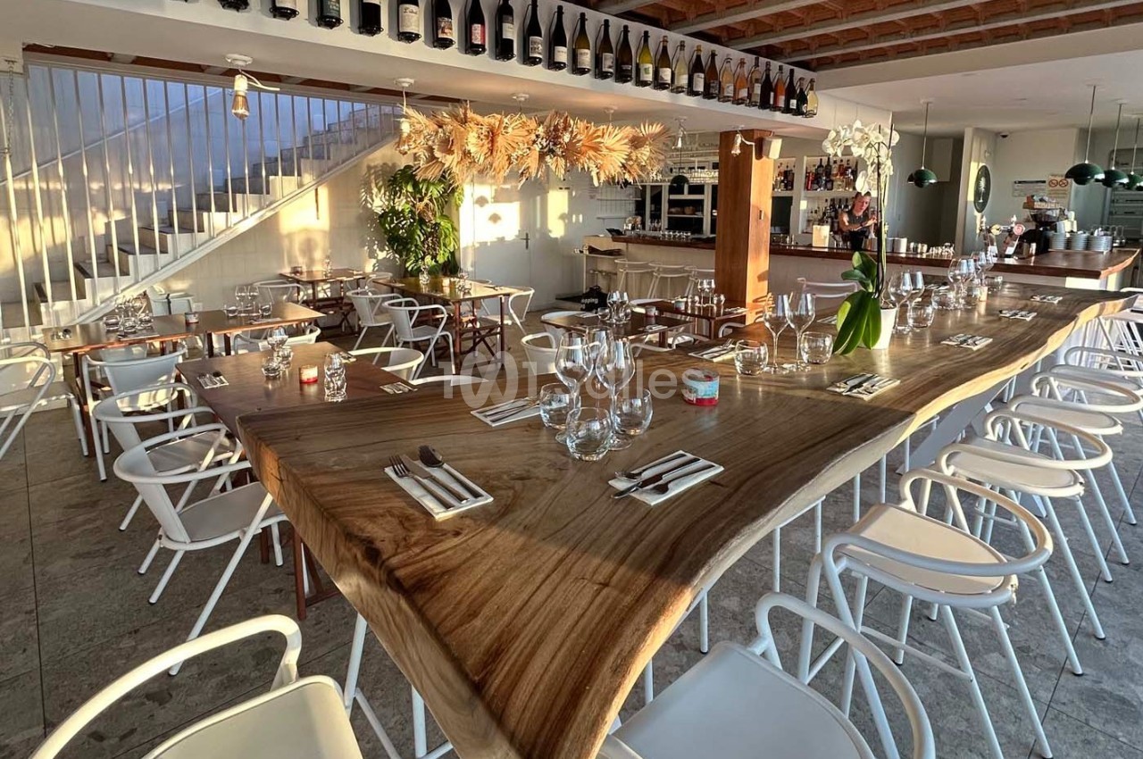 Salle de restaurant lumineuse avec une grande table en bois naturel, chaises blanches et décoration suspendue.