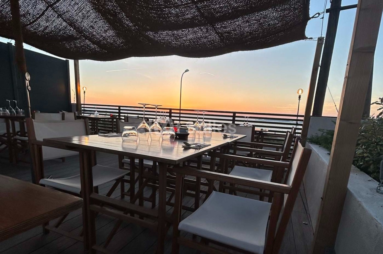 Table dressée en terrasse avec vue sur un coucher de soleil, chaises en bois et lumière tamisée.