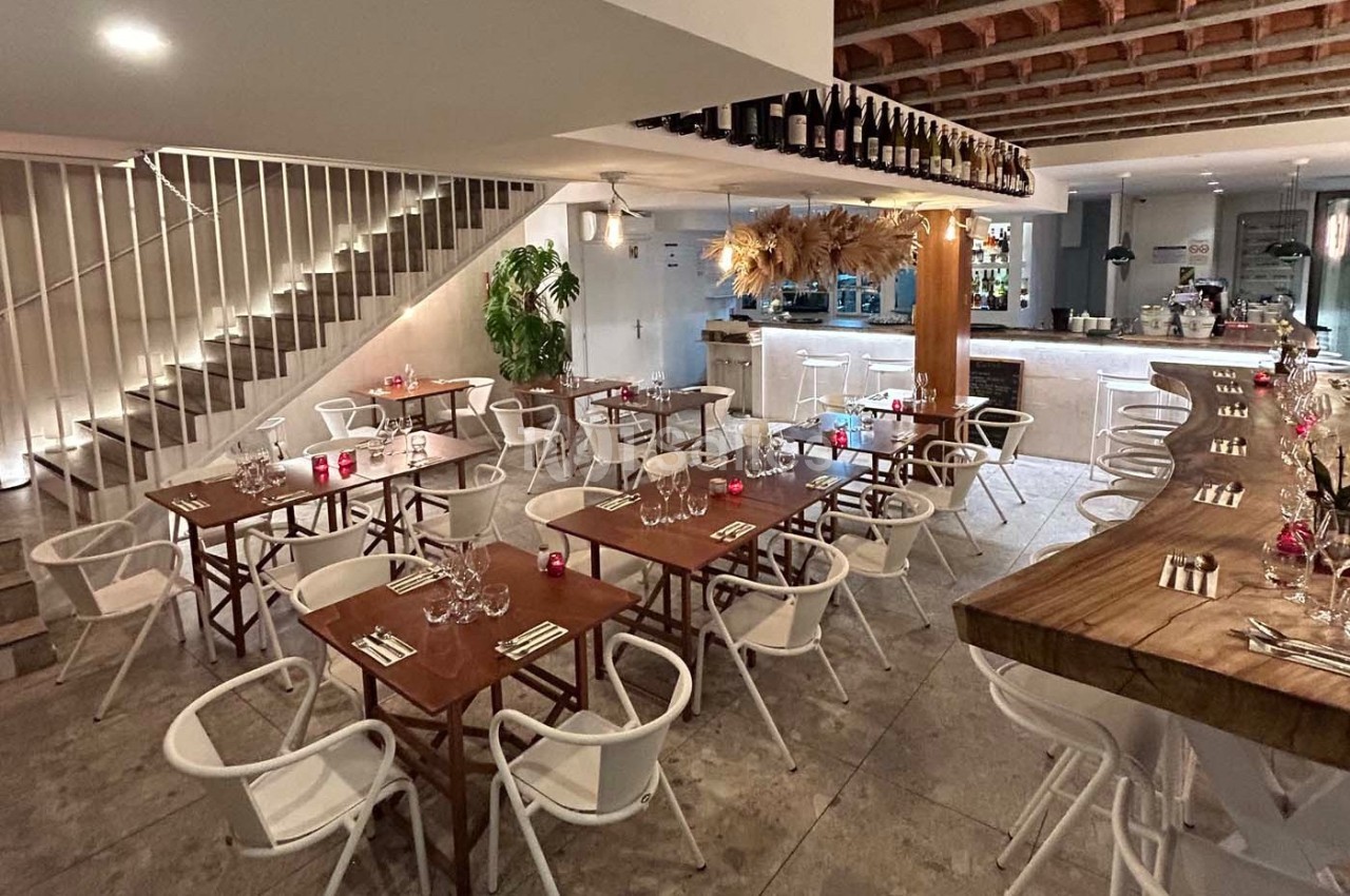 Salle de restaurant moderne avec tables en bois, chaises blanches, bar en arrière-plan et décoration suspendue.