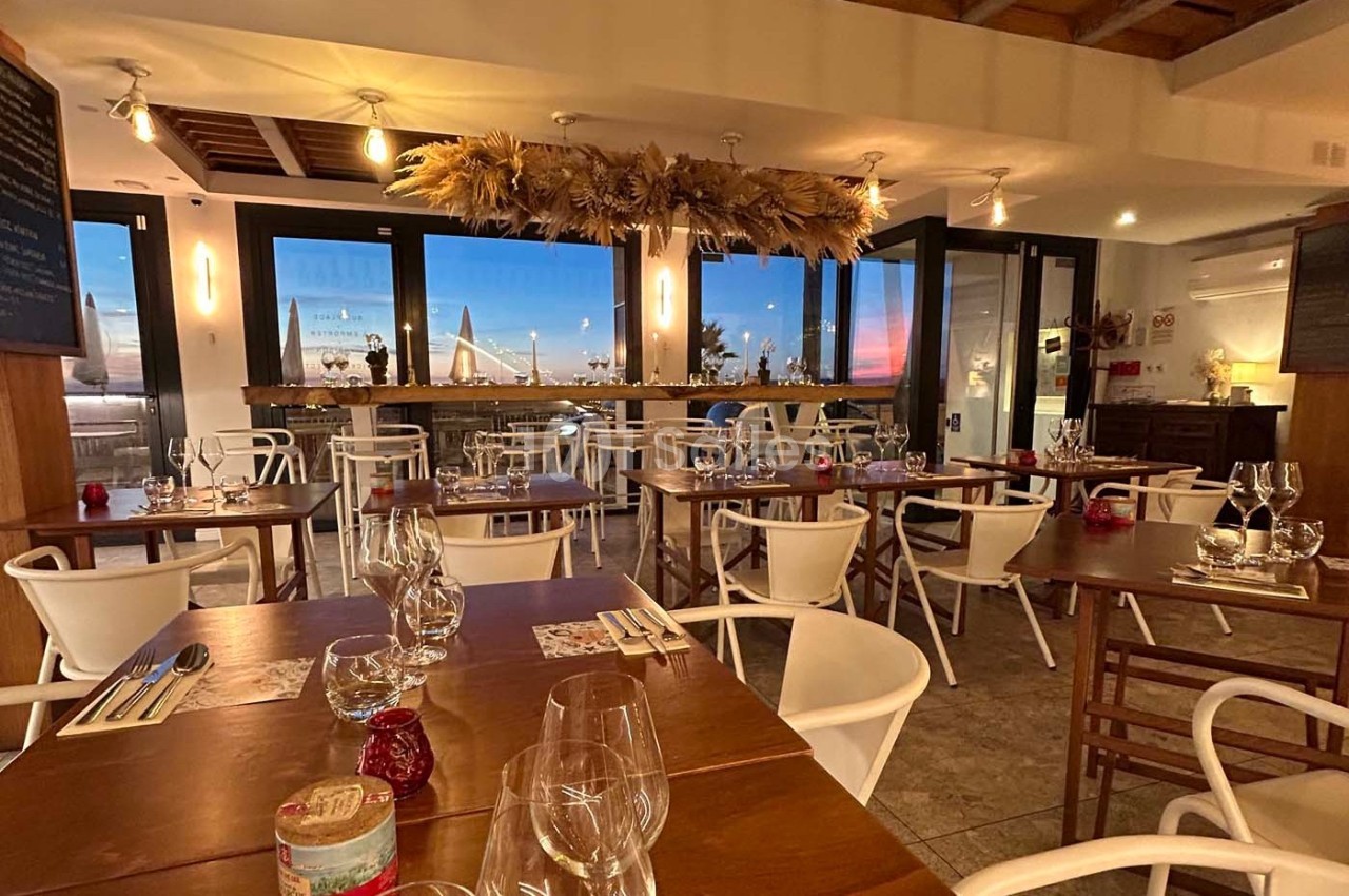 Salle de restaurant chaleureuse avec tables dressées, vue sur un pont illuminé à travers de grandes baies vitrées.