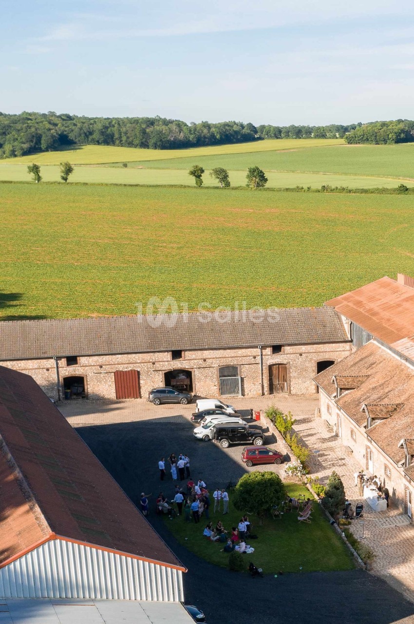 Vue aérienne d'une ferme en pierre entourée de champs verdoyants, avec des personnes rassemblées dans la cour.