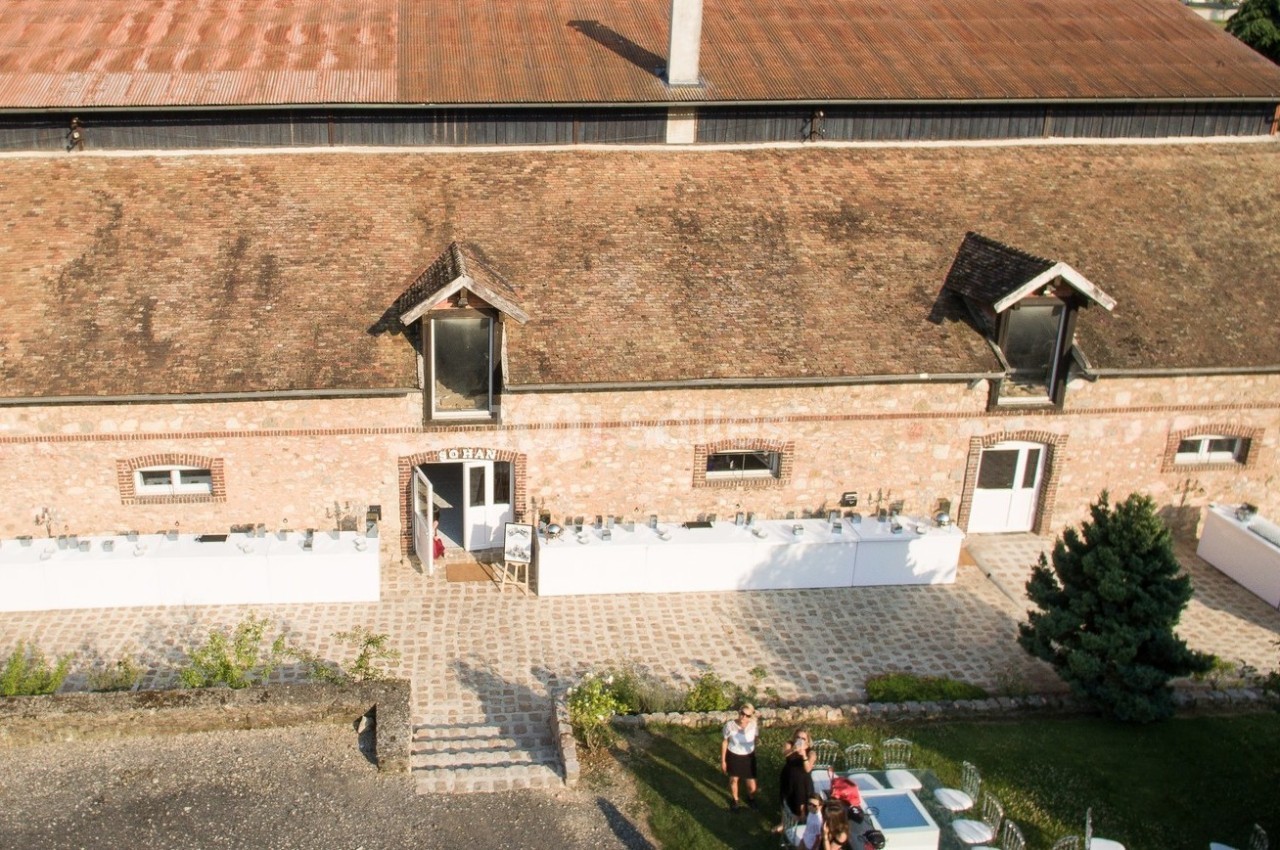 Bâtiment en briques avec toit en tuiles, petites fenêtres sous les combles, et terrasse aménagée avec des personnes.