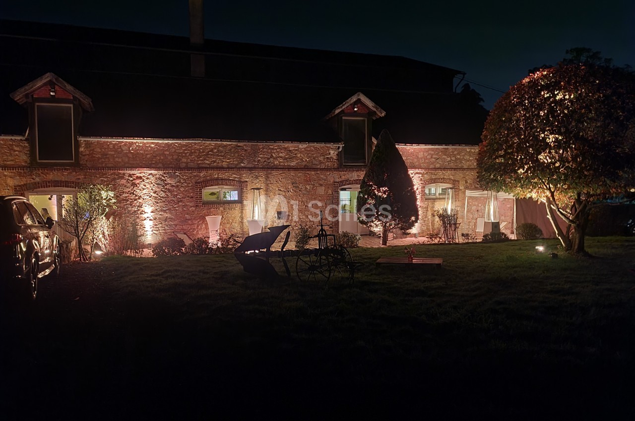 Façade en pierre d'une maison éclairée la nuit, entourée d'arbres, de mobilier de jardin et d'une pelouse.