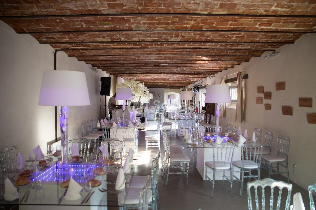 Salle de réception décorée avec des tables en verre, chaises transparentes et éclairage tamisé sous un plafond en briques.