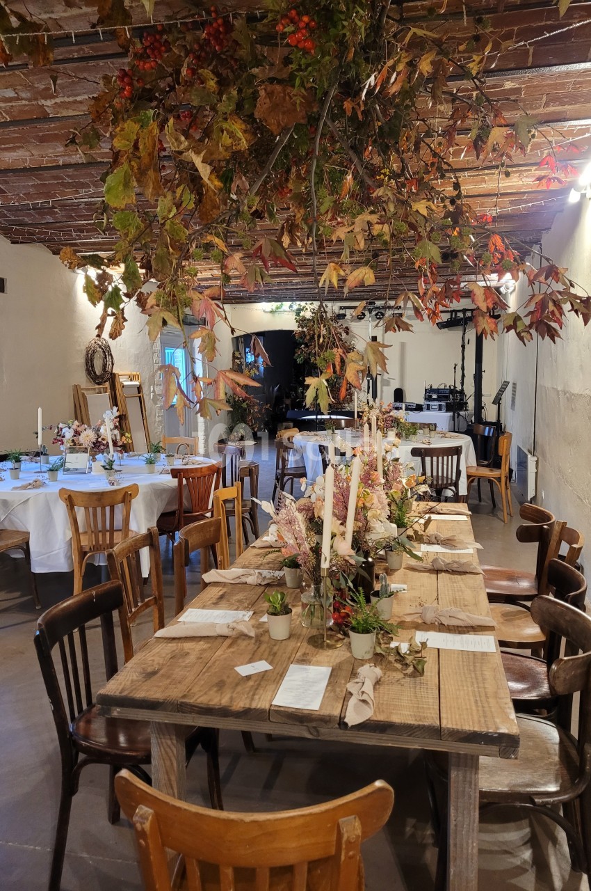 Salle décorée pour un événement, avec une grande table en bois ornée de bougies et de fleurs, sous un plafond rustique.