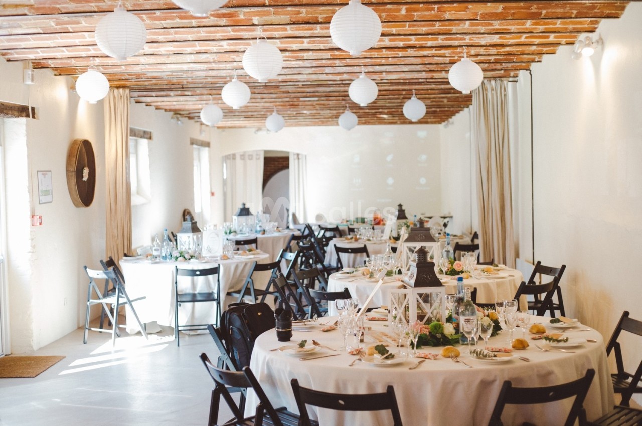 Salle de réception décorée avec des tables rondes, nappes blanches, chaises noires et lanternes suspendues au plafond.