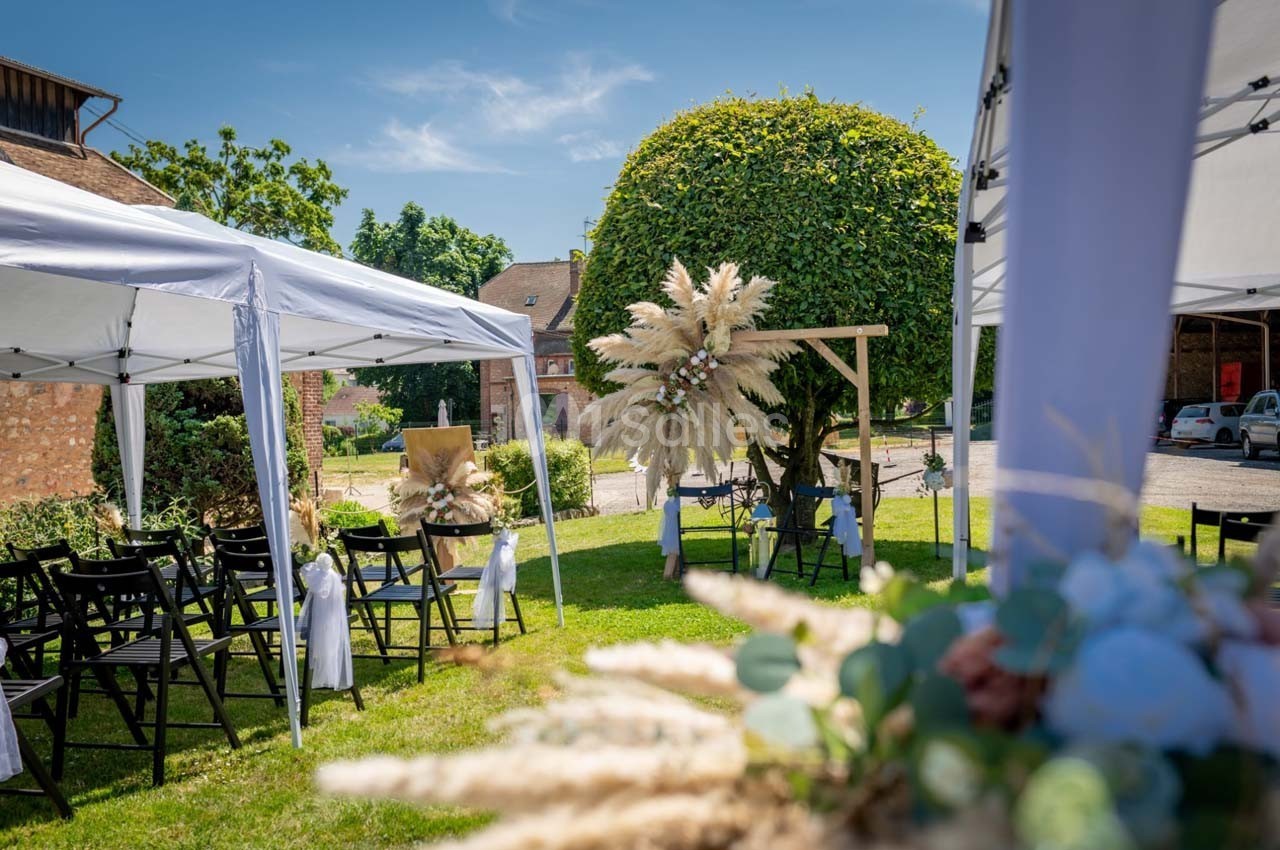 Cérémonie en plein air avec chaises noires, tonnelles blanches et décorations florales sous un arbre taillé.