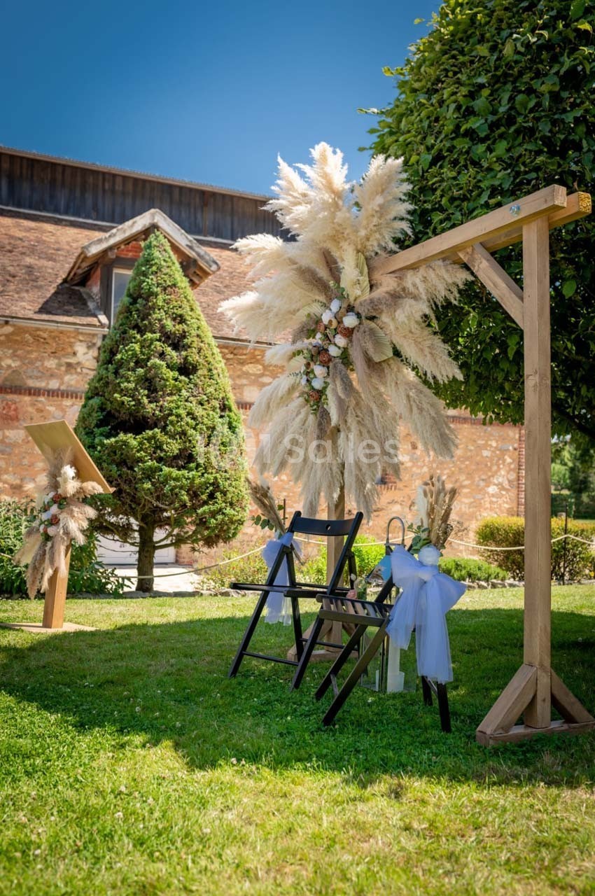 Décoration de cérémonie en extérieur avec arche en bois ornée de fleurs et de pampas, chaises noires décorées de rubans…