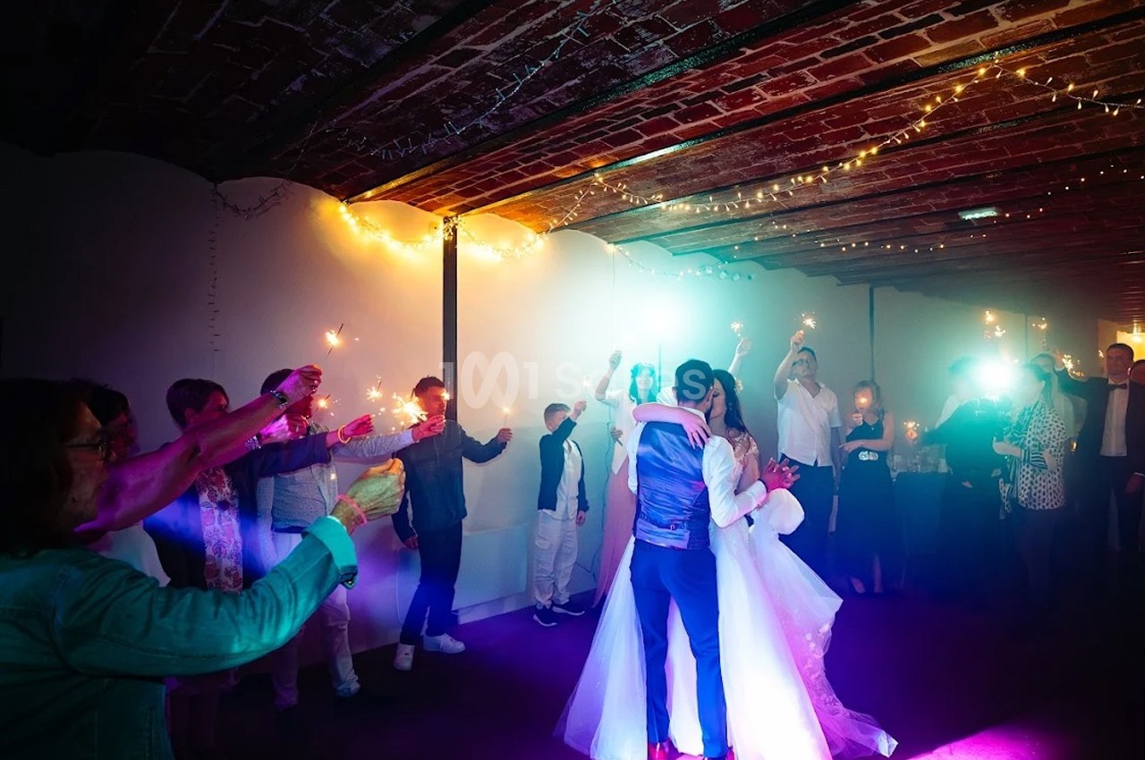 Un couple danse sous des lumières colorées, entouré d'invités tenant des cierges magiques dans une salle voûtée.