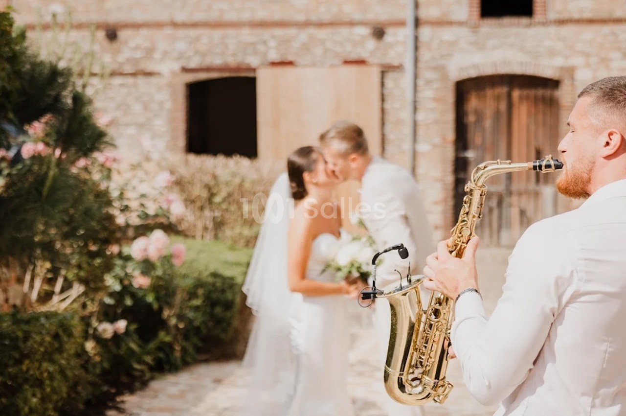 Un saxophoniste joue en extérieur tandis qu'un couple de mariés s'embrasse devant un bâtiment en pierre.