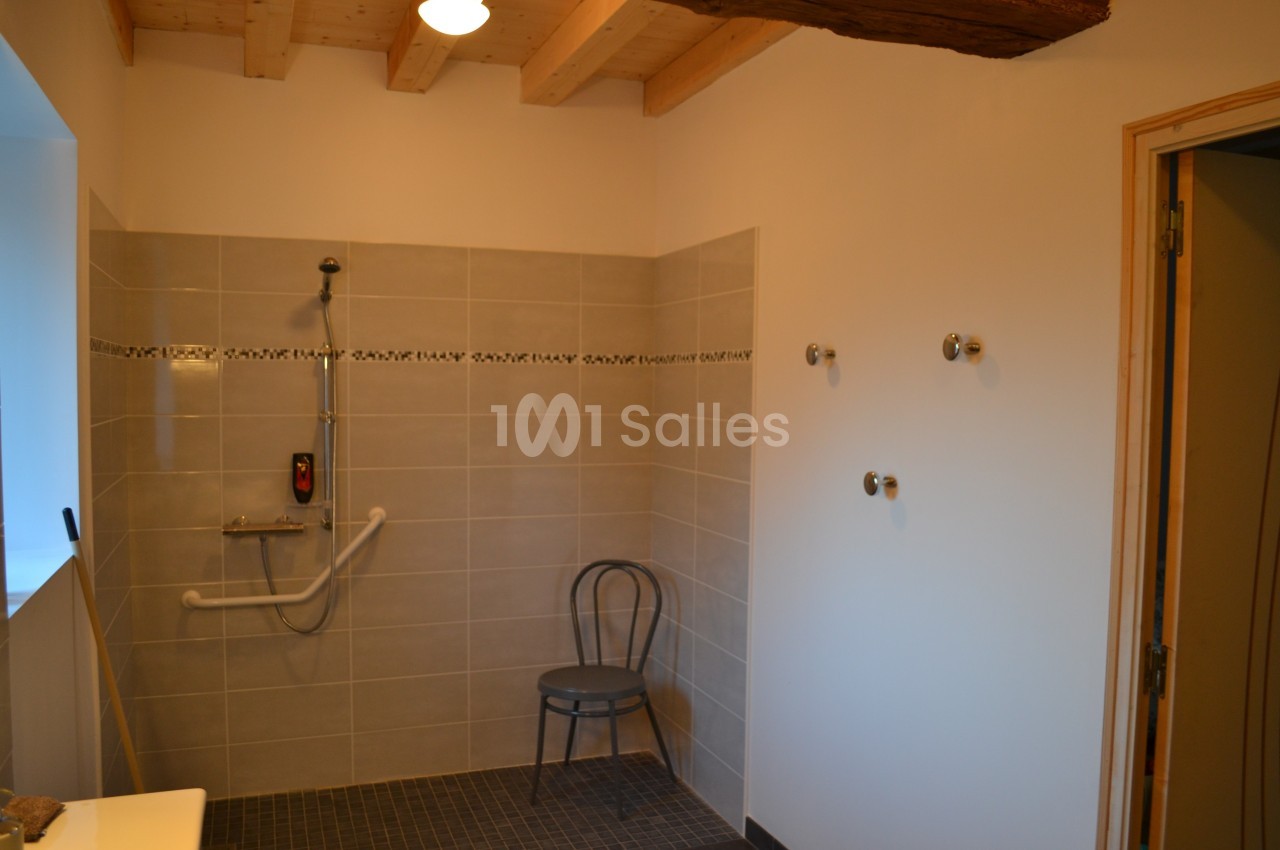 Salle de bain avec douche à l'italienne, barre d'appui, chaise et carrelage beige, éclairée par une lumière naturelle.