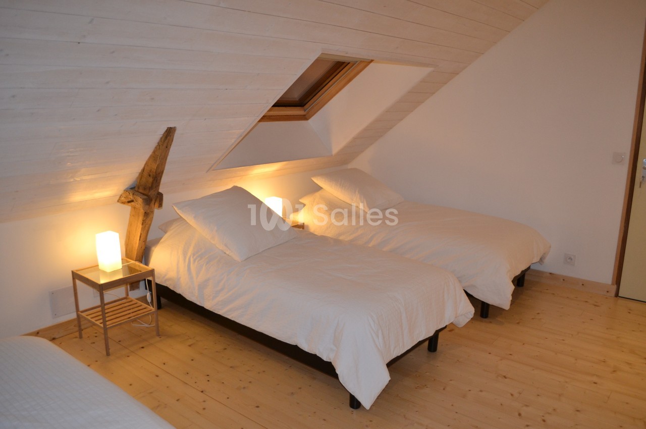 Trois lits simples avec draps blancs dans une chambre mansardée au plafond en bois clair, éclairée par des lampes de chevet.