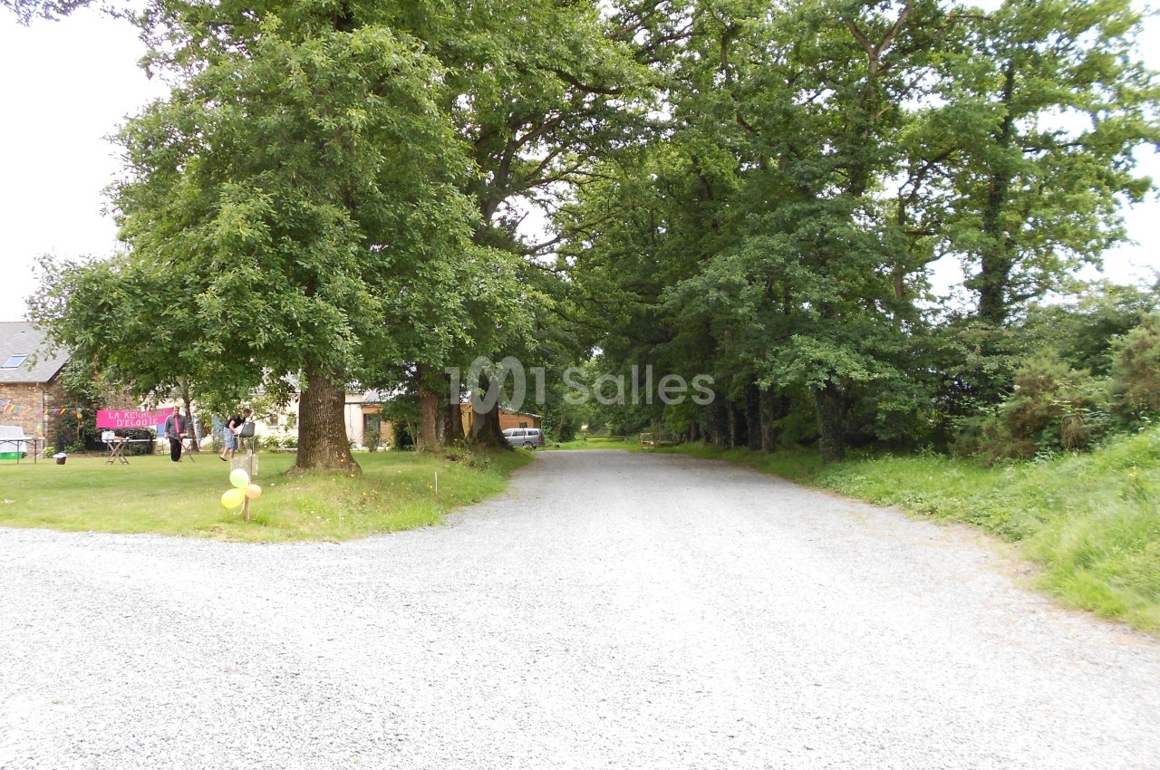 Chemin de gravier bordé d'arbres, avec des bâtiments et des personnes visibles sur la gauche.