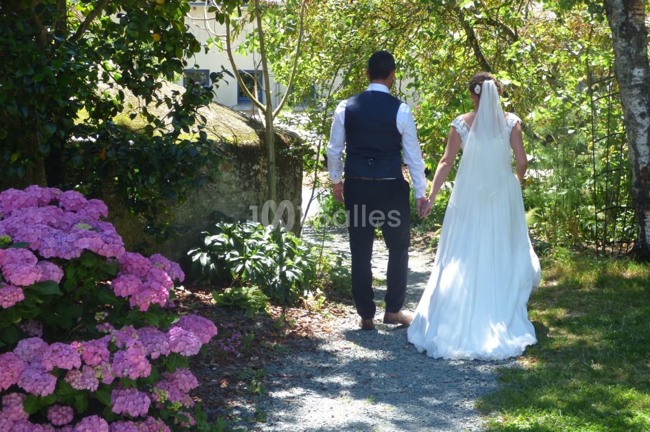Un couple en tenue de mariage marche main dans la main sur un chemin bordé d'hortensias et d'arbres.