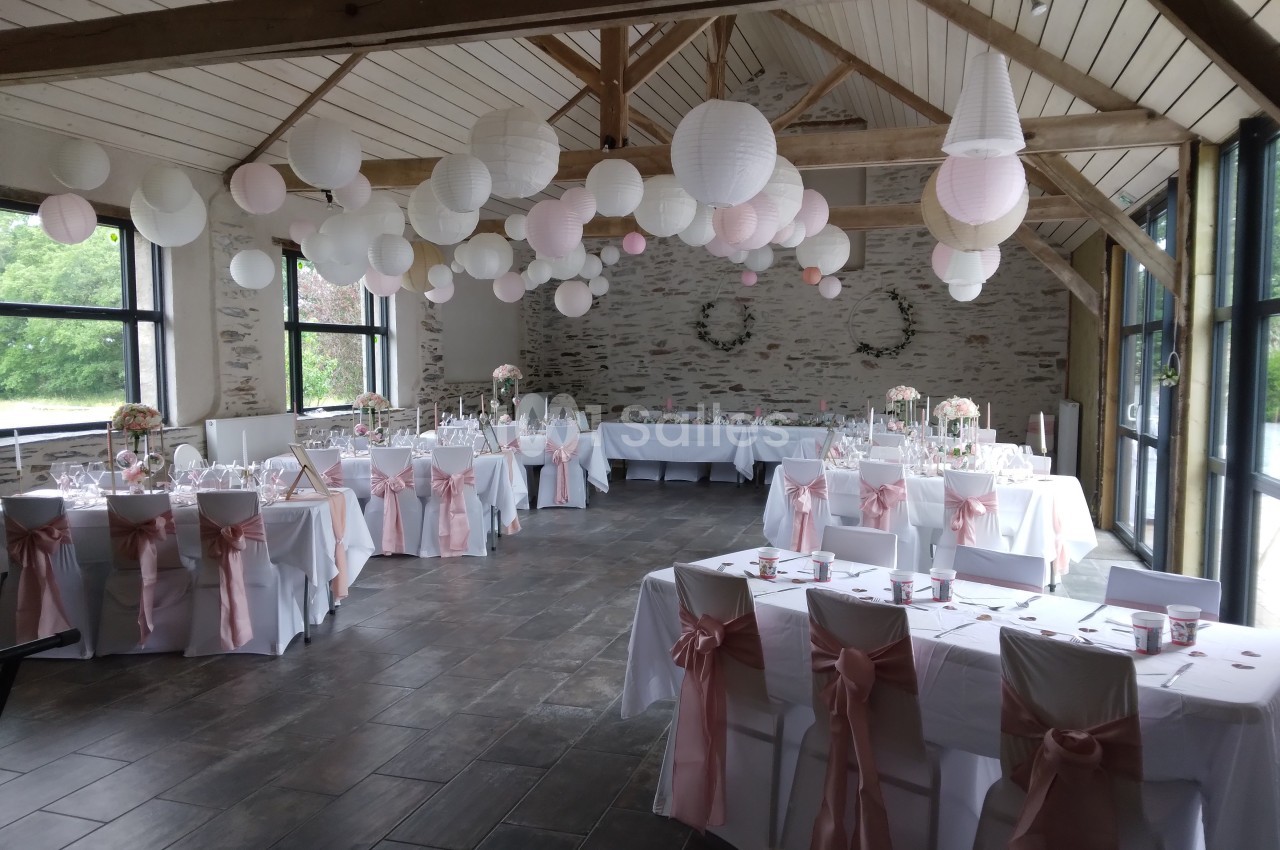 Salle de réception décorée pour un mariage avec des tables dressées, des chaises ornées de rubans roses et des lanternes…