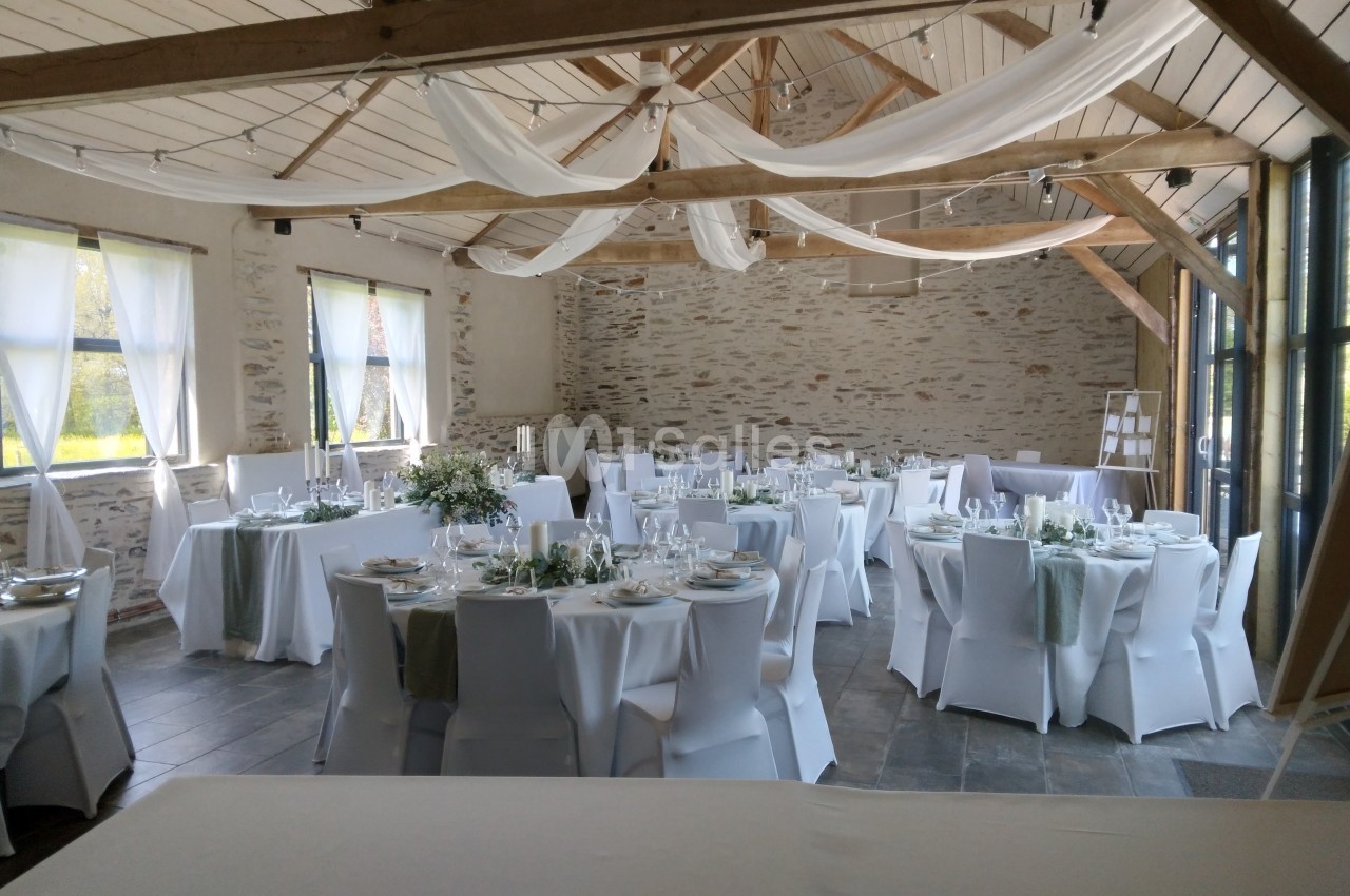 Salle de réception lumineuse avec tables rondes dressées, nappes blanches et décorations élégantes sous un plafond en bois.