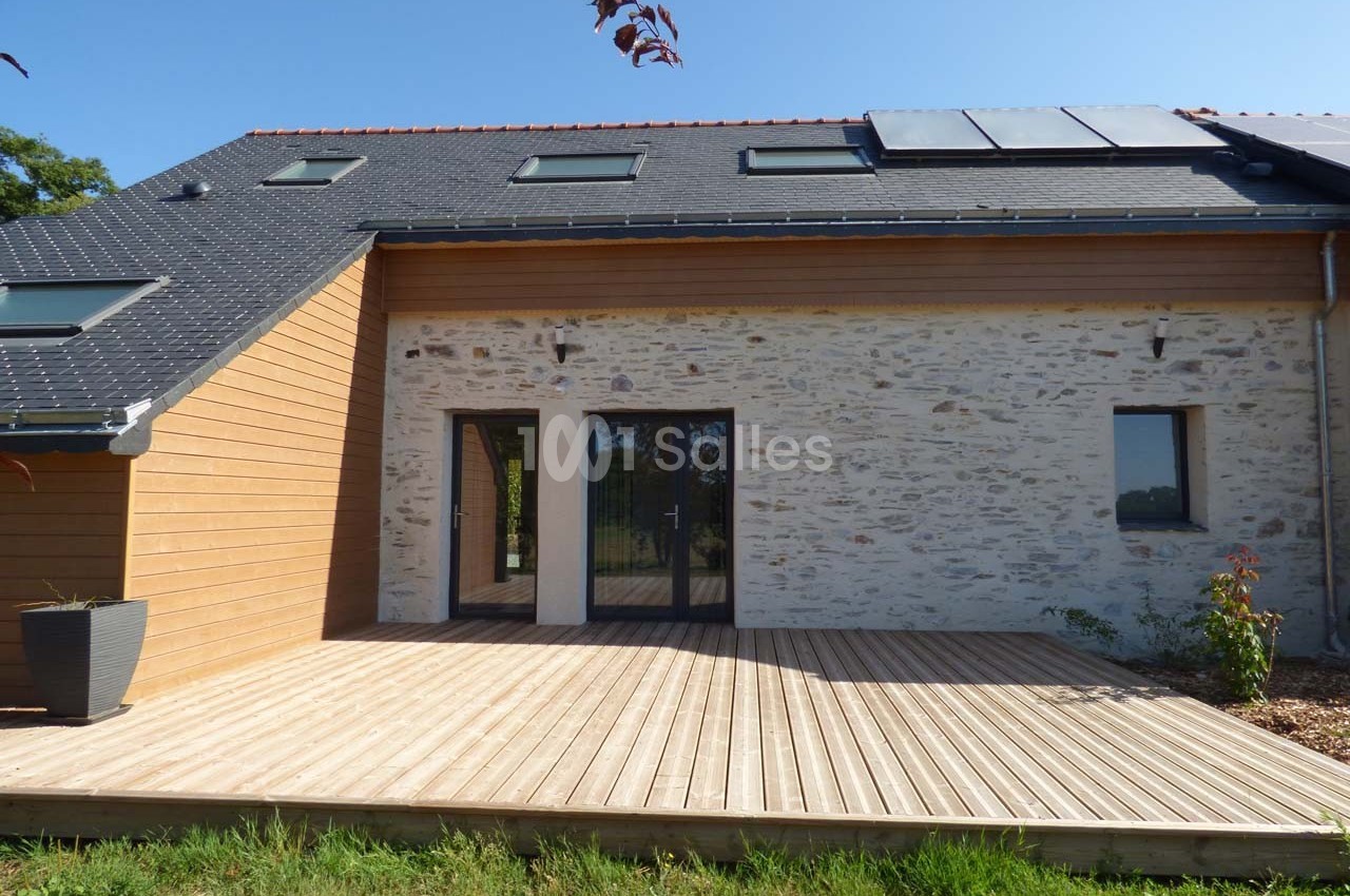 Façade d'une maison rénovée avec un mélange de bois et de pierre, terrasse en bois et panneaux solaires sur le toit.