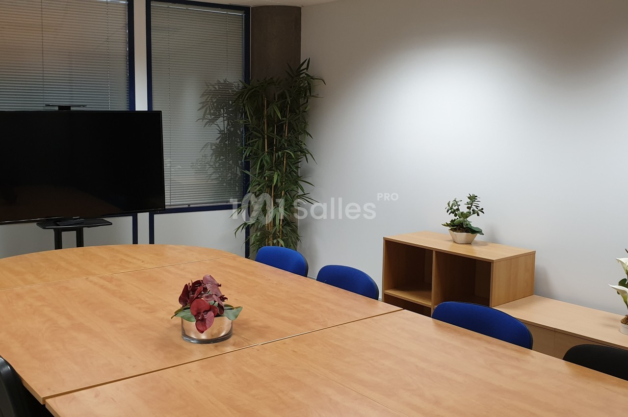 Salle de réunion avec grande table en bois, chaises bleues, écran, étagère et plantes décoratives.