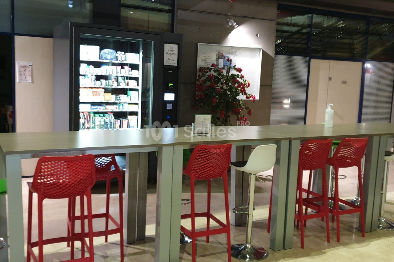 Table haute entourée de tabourets rouges et blancs, avec un distributeur automatique et une plante en arrière-plan.