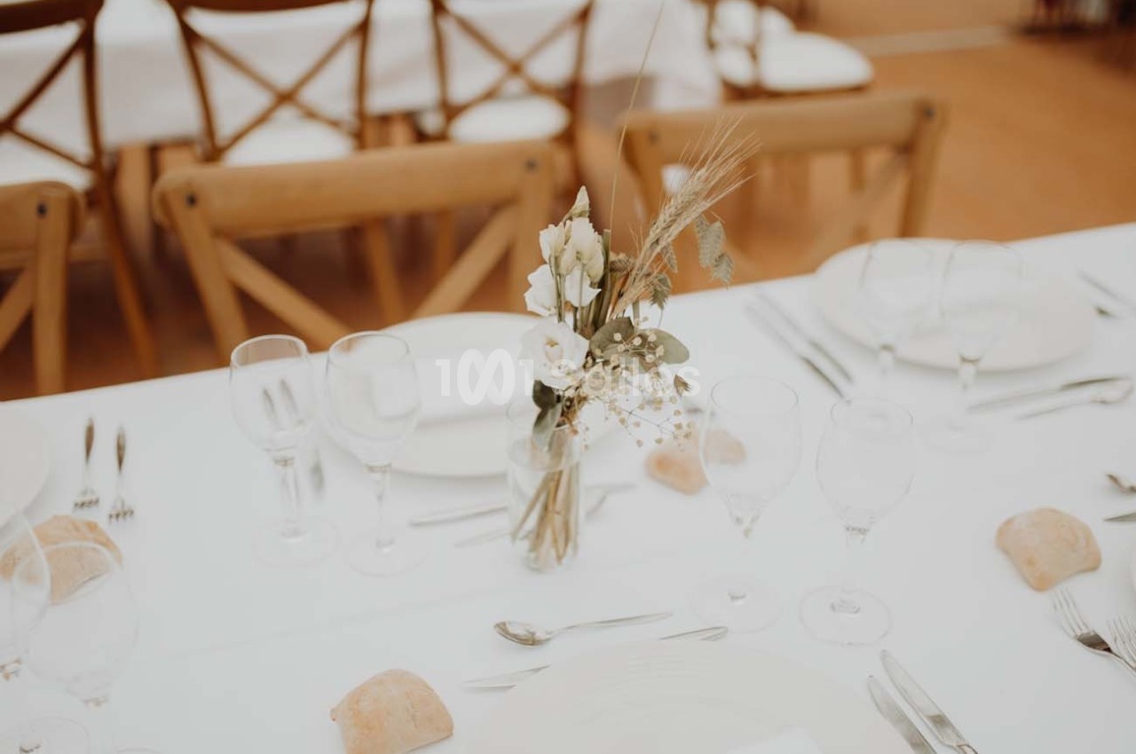 Table dressée avec nappes blanches, vaisselle, verres, couverts et un bouquet de fleurs séchées au centre.