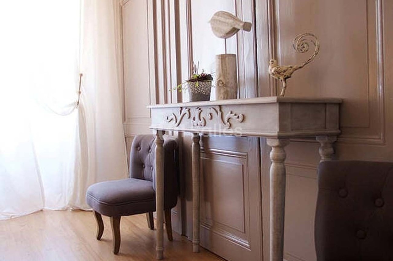 Console en bois sculpté avec décorations, chaise capitonnée grise et rideaux blancs dans une pièce lumineuse.