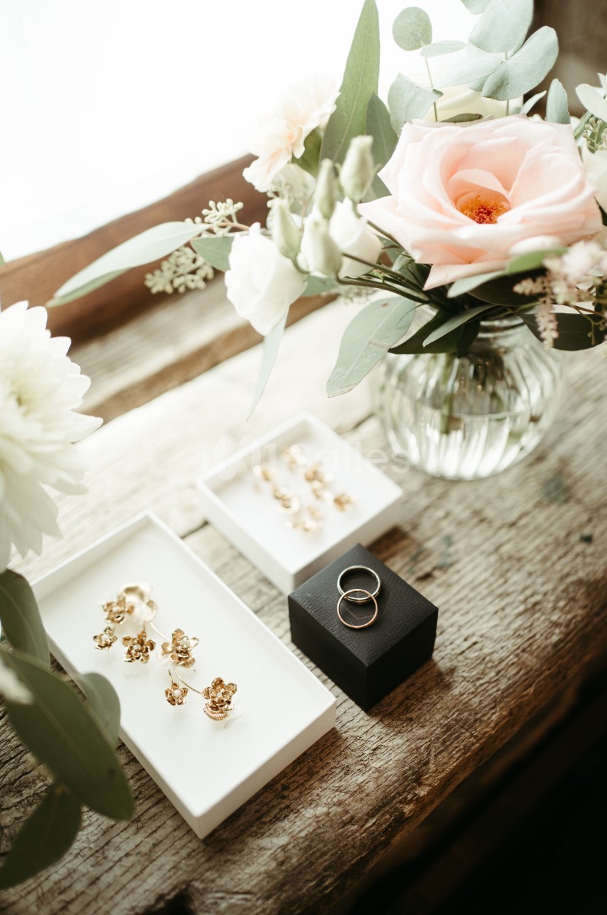 Boucles d'oreilles dorées et alliances posées sur une table en bois près d'un vase de fleurs.