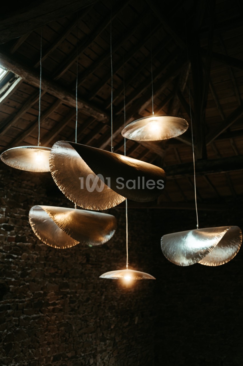 Suspensions lumineuses en métal aux formes organiques, accrochées sous un plafond en bois dans un espace en pierre.
