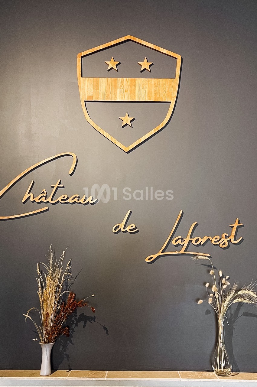Logo en bois du Château de Laforest sur un mur sombre, entouré de deux vases avec des compositions végétales.