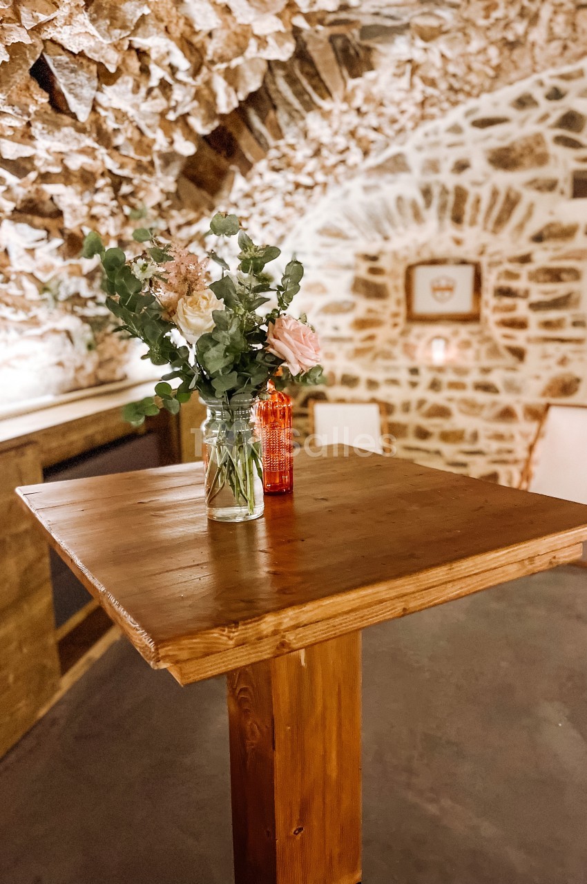 Table haute en bois avec un vase de fleurs et une bouteille rouge, dans une pièce aux murs en pierre apparente.